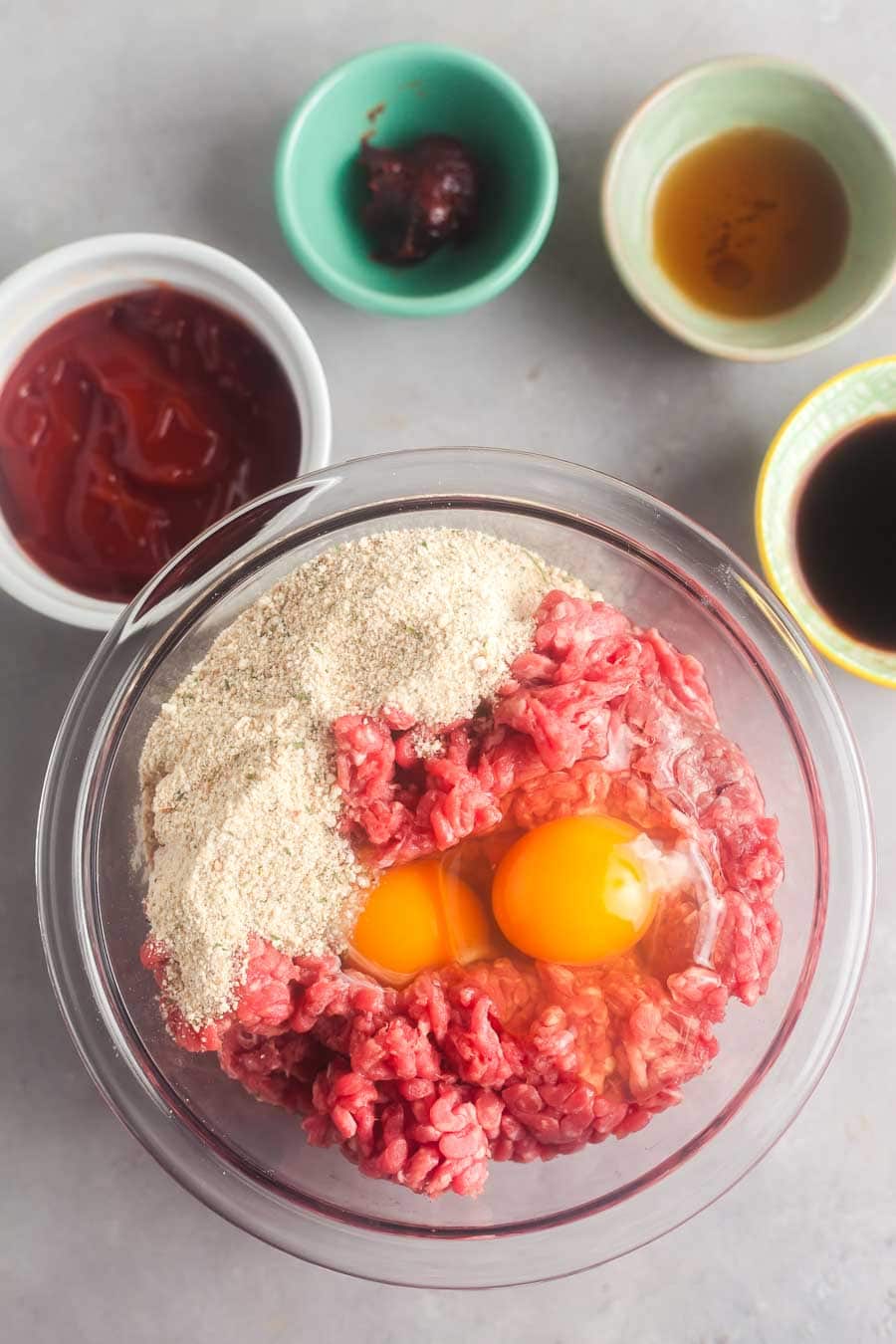 An overhead view of the ingredients to make slow cooker meatloaf. Ground beef, eggs, and breadcrumbs are together in a large bowl, The sauce ingredients are in individual bowls alongside